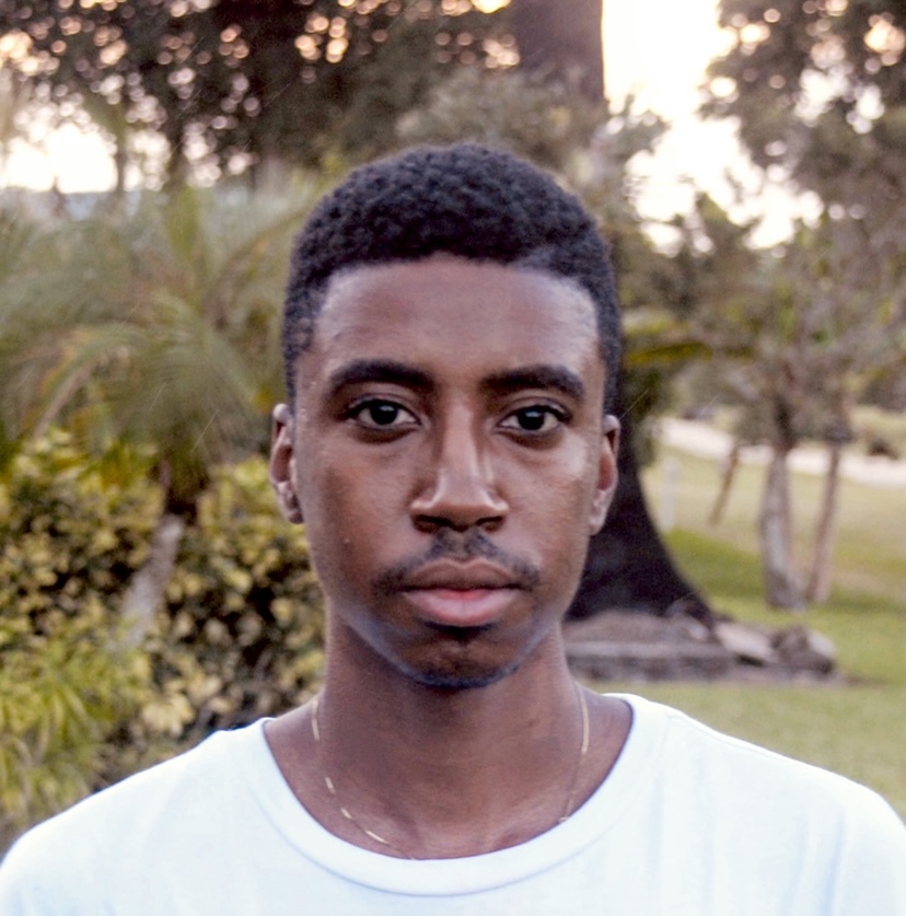 Portrait of Shonari Phillips outdoors at dusk, looking at the camera, with short black hair, a light mustache and a white T-shirt, trees and shrubs behind.