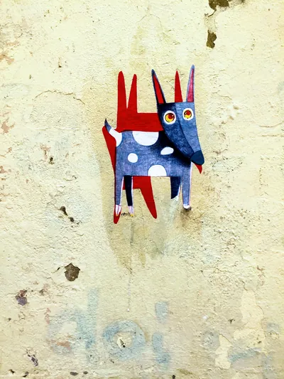 Blue and red cut-paper creature with polka dots stuck to a weathered cream wall.