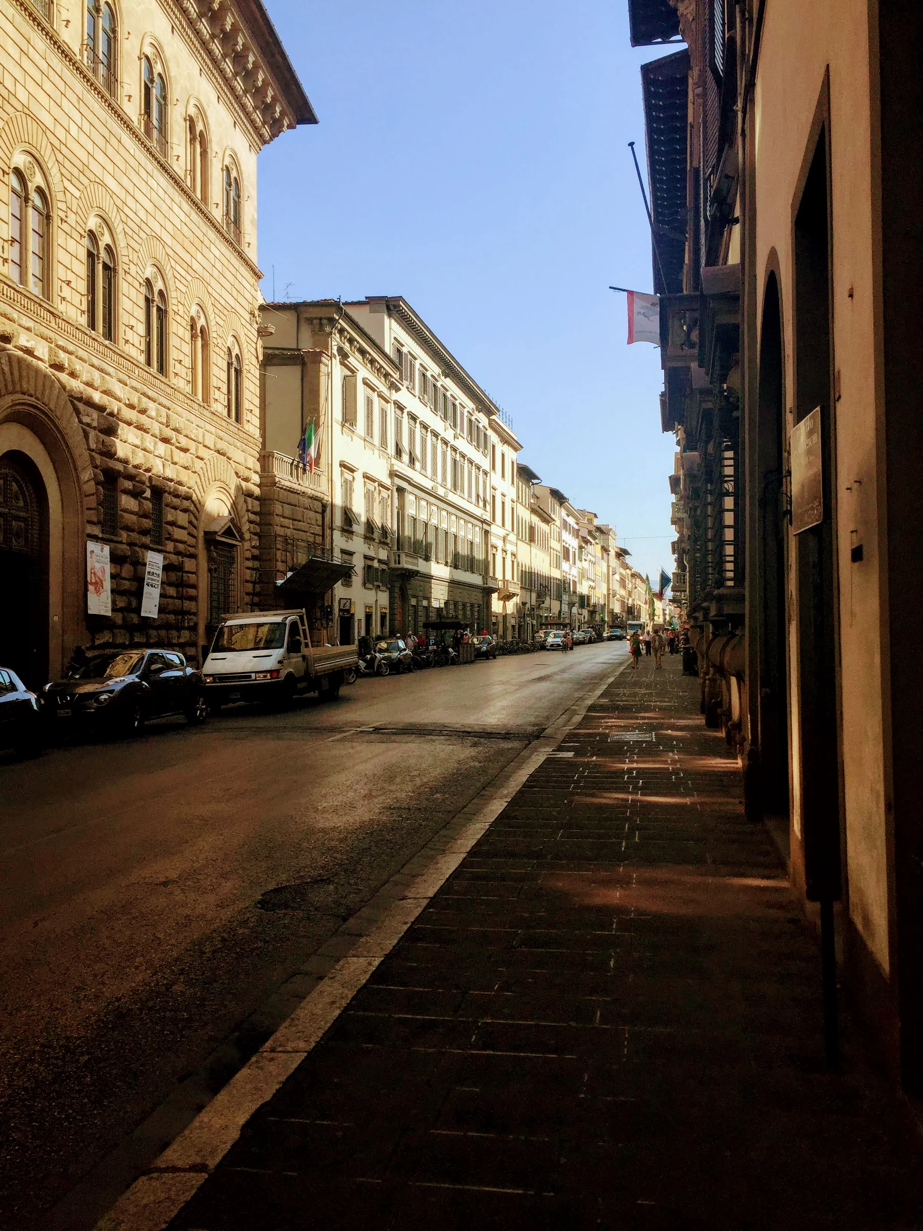 Sunlit city street with warm stone facades, parked cars and long shadows under a blue sky.