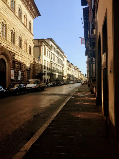 Sunlit city street with warm stone facades, parked cars and long shadows under a blue sky.