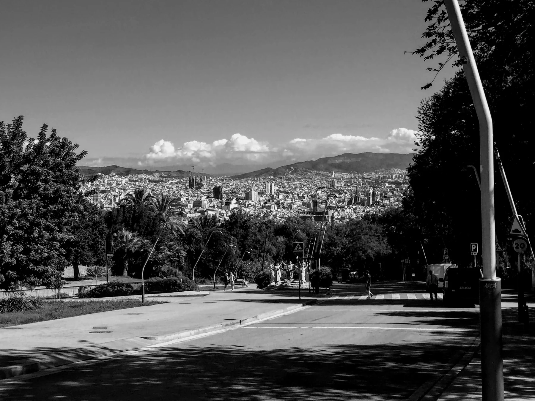 Black-and-white view from a tree-lined road over a sprawling city toward distant hills.