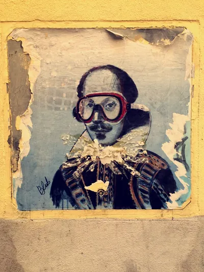 Street paste-up of Shakespeare wearing red swim goggles, peeling on a warm yellow wall.
