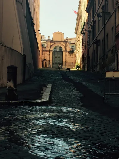 Wet cobblestone lane in cool shadow leading to a sunlit ochre church facade at the end.