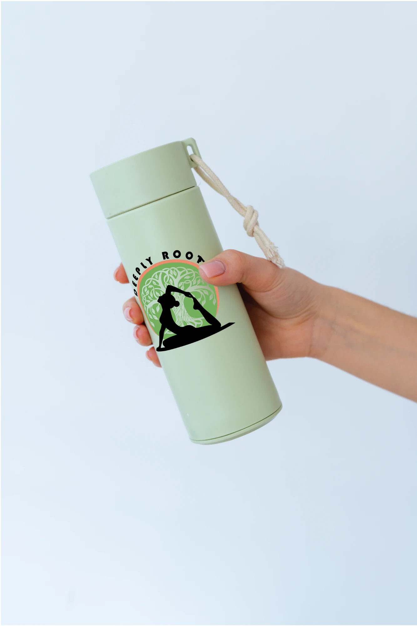 Hand holding a pale green bottle printed with the Deeply Rooted logo of a yoga pose in front of a tree inside a green and coral circle.