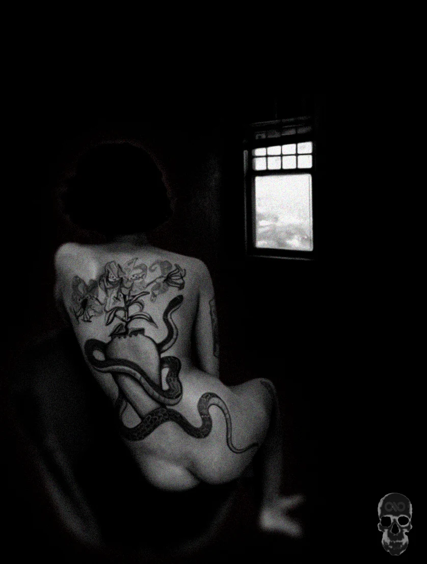 Black-and-white photo of a seated woman's tattooed back, a small bright window in the dark room.