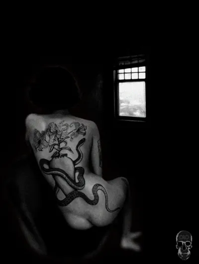 Black-and-white photo of a seated woman's tattooed back, a small bright window in the dark room.